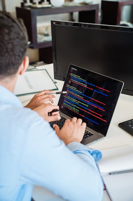 A person with dark hair and dressed in a light blue shirt is seated at a desk, working with a laptop that displays lines of colorful computer code. The desk contains a notebook and a desktop monitor p