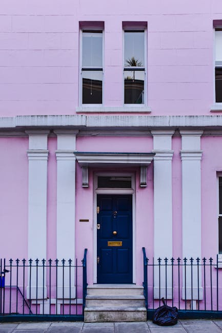 A close-up view of a pink residential building's entrance, featuring a dark blue wooden front door with a brass mail slot and door handle, accessed by three concrete steps with black metal handrails o